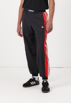 Osoba nosząca czarne spodnie dresowe Adidas z czerwonymi i białymi paskami po bokach oraz czarne buty sportowe Adidas z białymi paskami.