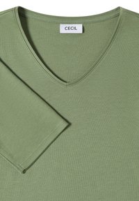 Chemise à manches longues vert clair en col en V avec couture visible et une étiquette blanche marquée "CECIL" au niveau du col.