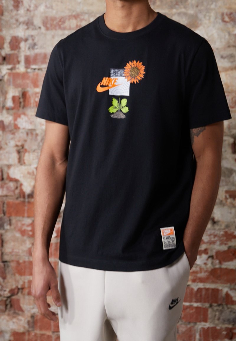 Person som bär en svart Nike T-shirt med en solrosgrafik och vita Nike-byxor, stående mot en bakgrund av tegelvägg.