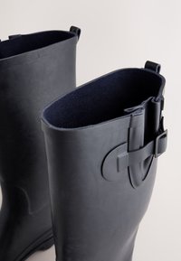 Schwarze Gummistiefel mit glatter Außenfläche, weichem marineblauem Innenfutter und einer Schnallendetail an der Seite für eine verstellbare Passform. Abgerundete Oberseiten.