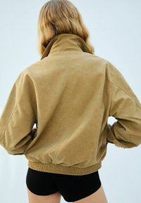 Beige, übergroße Jacke aus weichem Stoff mit hohem Kragen und elastischen Bündchen. Hat einen gerafften Saum und eine glatte Textur für besonderen Komfort.