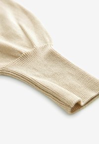 Beige neulekäsi, jossa on teksturoitu ribbikuvio ja joustava reunus. Tasapintainen kudos, jossa on hieno kiilto, kapeampi muoto ranteessa.