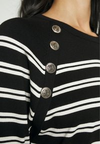Czarny i biały sweter w paski z okrągłym dekoltem, z ozdobnymi srebrnymi guzikami wzdłuż ramienia dla dodatkowego wykończenia.