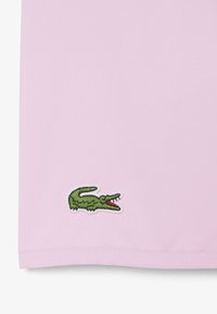 Tissu rose clair avec un petit logo de crocodile vert brodé près du bord inférieur.