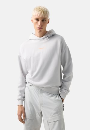 Jeune personne aux cheveux blonds courts portant un sweat à capuche gris clair avec le logo « FIRE+ICE » et un pantalon cargo assorti sur un fond uni.