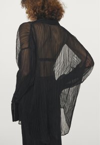 Schwarzes, plissiertes, durchscheinendes Hemd mit Kragen, langen Ärmeln und lockerer Passform. Hervorstechend durch den strukturierten Stoff und das fließende Design.