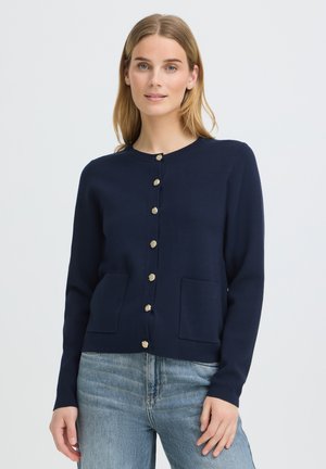 Femme aux cheveux blonds et raides portant un cardigan bleu marine boutonné avec deux poches et un jean bleu clair, debout devant un fond uni.