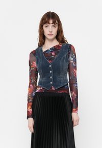 Denim veste ar V veida dekoltē, pieguļoša forma, pieci sudraba pogas un kontrastējošs izšuvums, kārtota pār ziedotu garām piedurknēm topu un melnu plisētu svārku.