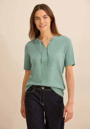 Camicia a maniche corte di un leggero color teal con scollatura a V e dettaglio con coulisse, realizzata in materiale morbido. Indossata con jeans in denim scuri, con cuciture visibili.