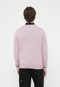 Svetlo roza pulover z teksturiranim pletenim vzorcem, ki ima črni ovratnik, rebraste manšete in pas. Hrbtni del je enobarven z zaobljenim izrezom na vratu.