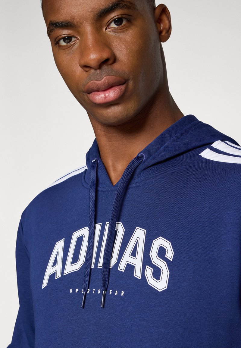 Navy blauwe hoodie van katoenmix, met een groot wit "ADIDAS" logo en sportieve witte strepen op de schouders.