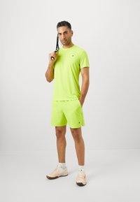 Hombre con un atuendo atlético de color verde neón sosteniendo una raqueta de tenis sobre su hombro, de pie frente a un fondo claro y liso.