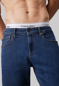 Jeans in denim blu scuro con un classico design a cinque tasche, zip a bottoni e un cinturino con logo Calvin Klein. Texture liscia con dettagli di cucitura.