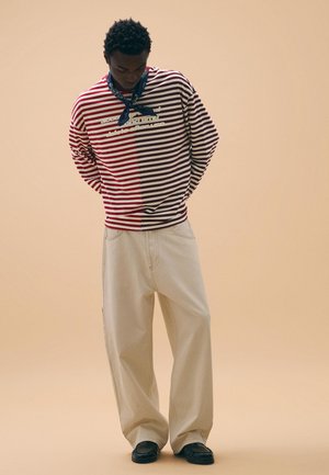 Moški v dvobarvnem rdeče-belih črtastih puloverju, bež hlačah širokih hlačnic, črnih čevljih in mornarsko modrem šalčku okoli vratu, stoji pred enobarvnim ozadjem.