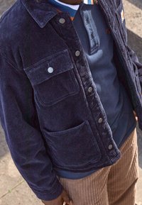 Giacca di velluto a coste blu navy con bottoni a pressione, due tasche a toppa e un colletto classico, indossata sopra una polo blu navy e pantaloni marroni con texture.