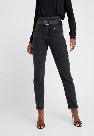 Frau trägt hoch taillierte schwarze Jeans mit einem breiten schwarzen Gürtel und schwarzen High-Heeled Sandalen mit Riemchen, steht vor einem schlichten Hintergrund.