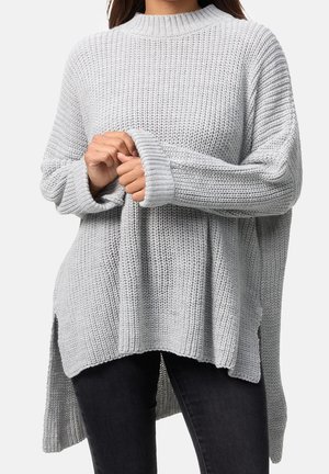 Personne portant un pull en maille ample gris clair à manches longues, tenant une manche avec les deux mains, associé à un pantalon foncé.