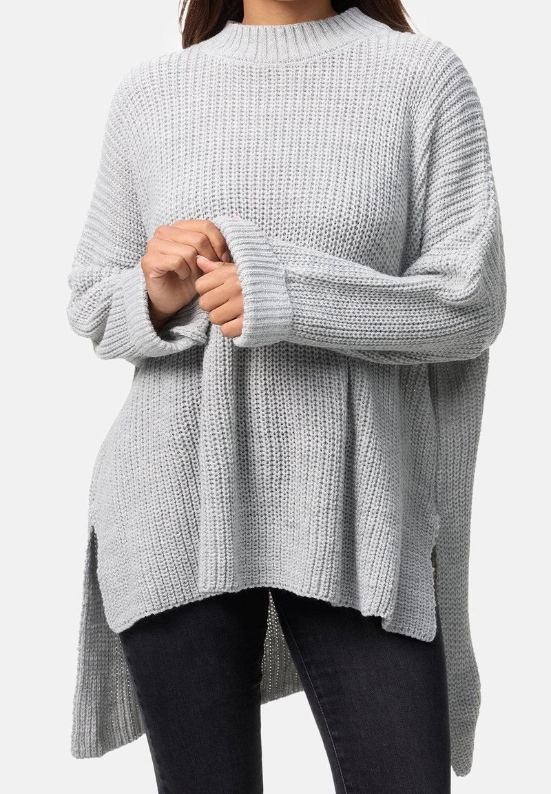 Personne portant un pull en maille ample gris clair à manches longues, tenant une manche avec les deux mains, associé à un pantalon foncé.