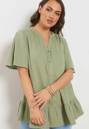 Kvinde iført en løs, lysegrøn knap-op bluse med en flæsekant, guldbøjler og lyseblå jeans, poserer med krydsede hænder.