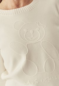 Pulover alb din tricot cu un design în relief reprezentând un ursuleț de pluș pe partea din față. Gulerul este ribatat, având o textură moale pe tot parcursul.