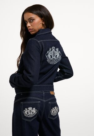 Kvinde med langt hår iført mørk denim Juicy Couture jakke og jeans med hvide broderede logoer på ryggen og lommerne, som kigger over skulderen.