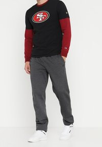 Camiseta negra con mangas largas rojas, logo de la NFL en el pecho, combinada con pantalones de chándal grises y zapatillas blancas. Tejido suave, corte casual.