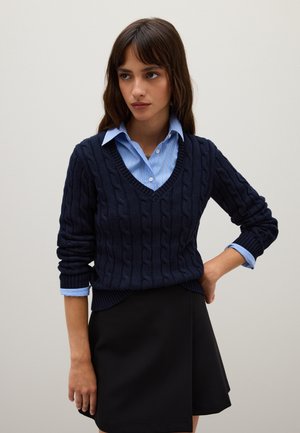 Joven mujer con un suéter de punto trenzado azul marino de cuello en V sobre una camisa de cuadros azul y una falda negra, de pie con una mano en la cadera.