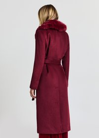 Langer burgunderroter Mantel mit Kunstpelzkragen, Taillengürtel und langen Ärmeln. Texturiertes Gewebe, mittellang bis zur Wade, minimalistische Details.