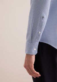 Camicia a quadri blu con polsino abbottonato, caratterizzata da un tessuto testurizzato e bottoni bianchi a contrasto. Braccio posizionato accanto a un tessuto scuro.