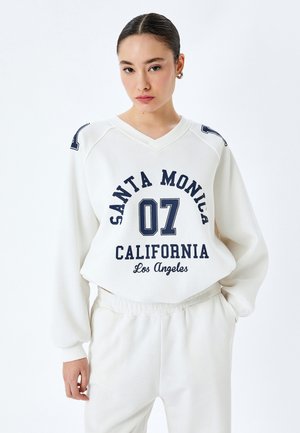 Hvid sweatshirt med marineblå print, der har "SANTA MONICA 07 CALIFORNIA" og "Los Angeles." V-hals, afslappet pasform og sænkede skuldre.