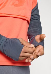 Αθλητική μπλούζα με μακριά μανίκια, που διαθέτει φωτεινό κοραλλί πάνω μέρος και σκούρους γκρι μανίκια. Το ύφασμα φαίνεται ελαφρύ με ανοίγματα για τους αντίχειρες.