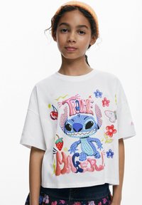 Vit kortärmad t-shirt med en färgglad grafisk bild av Stitch, omgiven av blommor, jordgubbar och ordet "TROUBLE" i lekfulla typsnitt.
