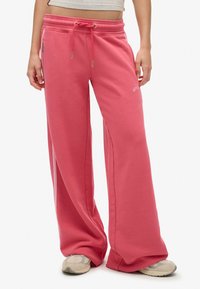 Persona che indossa pantaloni ampi corallo con vita regolabile e sneaker beige, in piedi contro uno sfondo bianco.