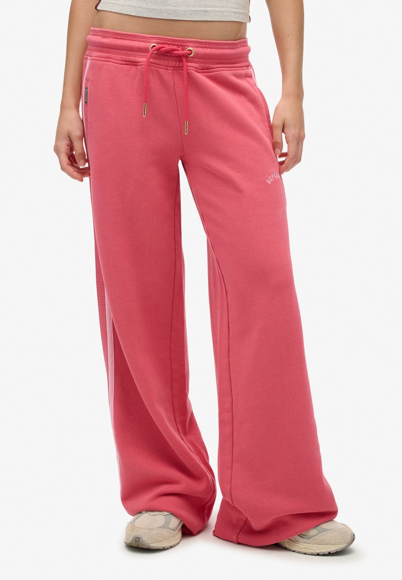Persona che indossa pantaloni ampi corallo con vita regolabile e sneaker beige, in piedi contro uno sfondo bianco.