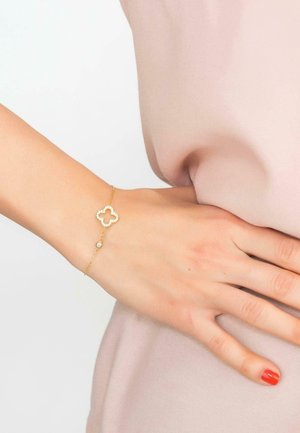 Hand with red nail polish wearing delicate gold bracelet with diamond-studded clover charm resting on light pink fabric.