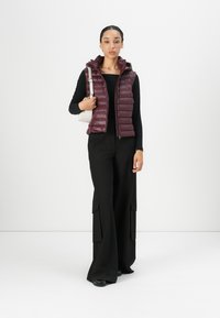 Burgunderfarget quilted puffer-vest med høy krage, båret over en svart langermet topp. Svart vide bukser og liten hvit skulderveske.