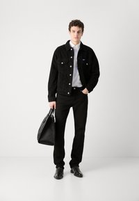 Veste en velours côtelé noire avec fermeture à boutons et deux poches avant, chemise blanche en dessous, pantalon noir et sac fourre-tout noir texturé.