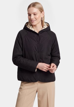 Femme aux cheveux blonds portant une veste noire à capuche avec une doublure à motif d'étoiles et un pantalon beige, souriante et regardant vers le bas.