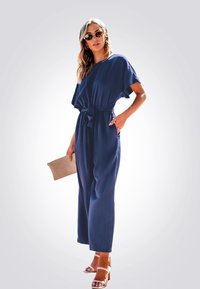 Combinaison bleu marine à coupe décontractée, manches courtes, taille ceinturée et jambes larges, associée à une pochette beige et des sandales de couleur claire.