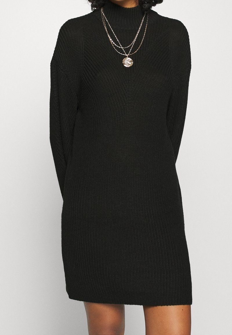 Svart stickad klänning med hög hals, långa ärmar och lös passform. Har en ribbad textur och slutar ovanför knäet. Lager av gyllene kedjor.