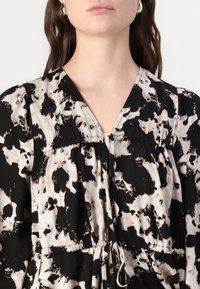 Frau in einem schwarz-weißen, abstrakt gemusterten Kleid mit V-Ausschnitt und Kordelzug in der Taille, trägt außerdem goldene Creolen.