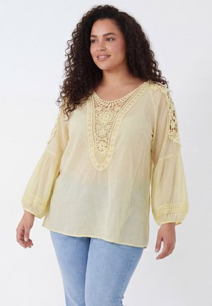 Vrouw met lang krullend haar lacht, draagt een losse bleekgele blouse met kanten details en lichtblauwe spijkerbroek tegen een witte achtergrond.
