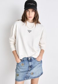 Biely sveter s logom "Tommy Jeans", skombinovaný s svetlomodrou džínsovou sukňou, na ktorej sú zaoblené vyšívané nášivky. Čierna čiapka dokončuje vzhľad.