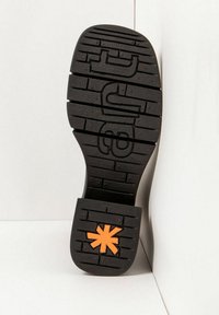 Suela de zapato negra con surcos rectangulares y un logotipo en forma de asterisco naranja cerca del talón, sobre un fondo blanco en una esquina.