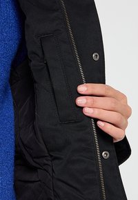 Main tenant une veste noire ouverte montrant une poche intérieure et une fermeture éclair argentée, avec un tissu bleu visible sur le côté gauche.