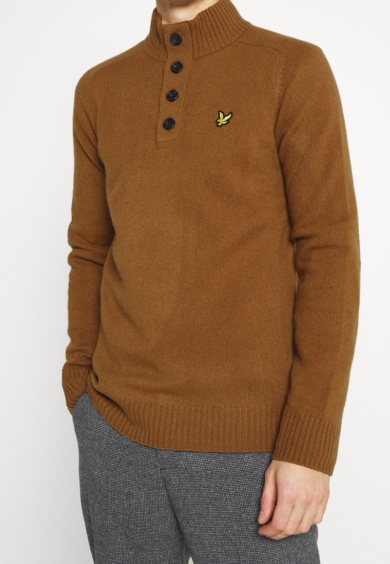 Bruine gebreide trui met een hoge geribbelde kraag, vijf zwarte knopen en een klein geel logo. Heeft een aansluitend ontwerp en geribbelde manchetten.