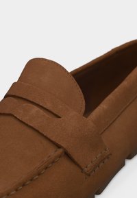 Mocassin en daim marron avec bride à monnaie, coutures détaillées et petit talon, présenté en gros plan sur fond blanc.
