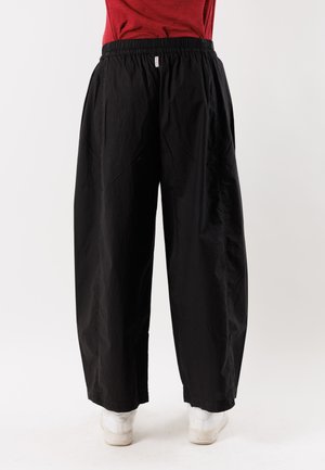 Persona che indossa pantaloni neri larghi con vita elastica e scarpe bianche, in piedi contro uno sfondo chiaro e uniforme.