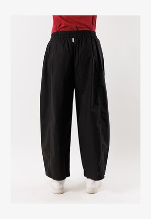 Persona che indossa pantaloni neri larghi con vita elastica e scarpe bianche, in piedi contro uno sfondo chiaro e uniforme.