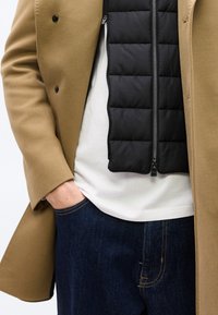 Άνδρας που φοράει παλτό καμηλοπάρδαλης πάνω από μαύρο ενισχυμένο γιλέκο και λευκό πουκάμισο, με το χέρι στην τσέπη σκούρων μπλε τζιν.